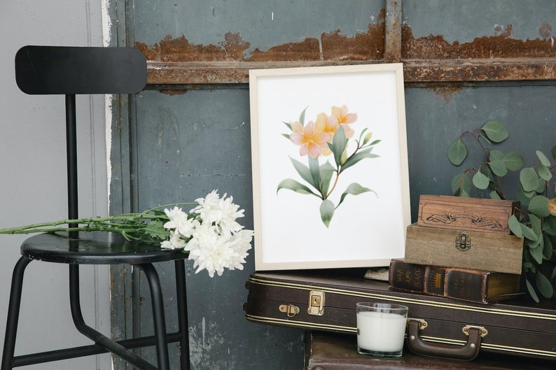 May include: A framed watercolor print featuring a floral arrangement with yellow and pink blossoms and green leaves. The artwork is displayed on a white background. The image also includes a black stool, white flowers, and vintage suitcases.