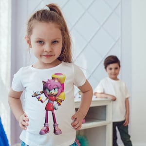 May include: A young girl wearing a white t-shirt with a pink and yellow cartoon character holding a hammer. The character is wearing a red dress and white boots. The girl is smiling and looking at the camera.