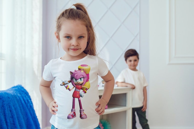 May include: A young girl wearing a white t-shirt with a pink and yellow cartoon character holding a hammer. The character is wearing a red dress and white boots. The girl is smiling and looking at the camera.
