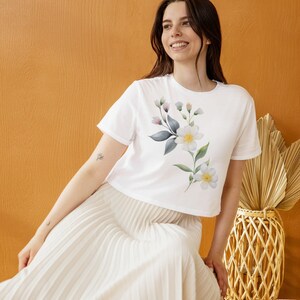 May include: White crop top with a floral design featuring white flowers, green leaves, and small purple buds. The model is wearing a white pleated skirt. The background is a solid orange color.