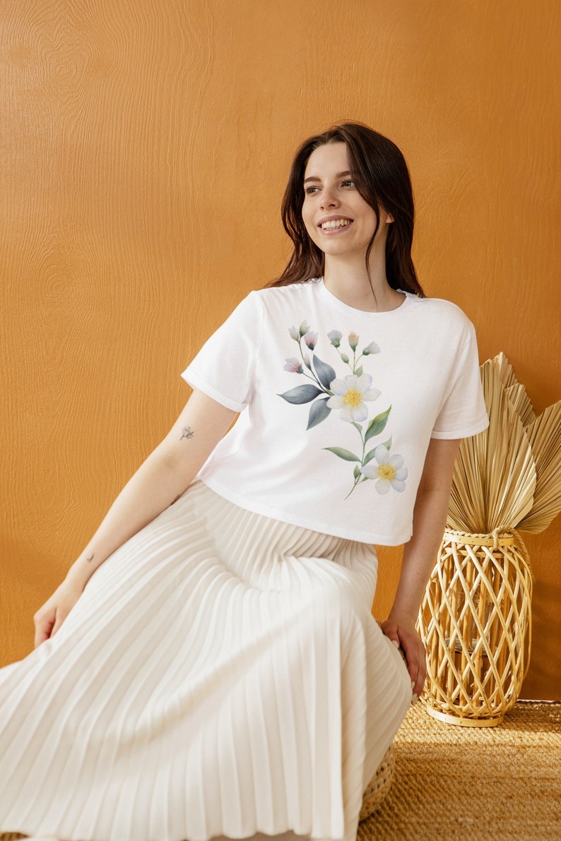 May include: White crop top with a floral design featuring white flowers, green leaves, and small purple buds. The model is wearing a white pleated skirt. The background is a solid orange color.