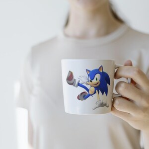 May include: White ceramic mug with a blue and red cartoon character of Sonic the Hedgehog running. The character is in a dynamic pose with his arms and legs outstretched.