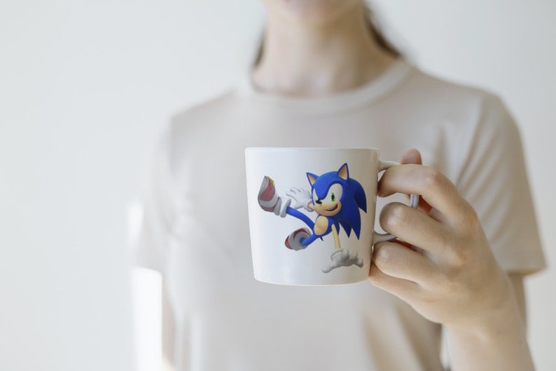 May include: White ceramic mug with a blue and red cartoon character of Sonic the Hedgehog running. The character is in a dynamic pose with his arms and legs outstretched.