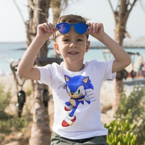 May include: A young child wearing a white t-shirt with a blue Sonic the Hedgehog graphic. The child is wearing blue sunglasses and is smiling.