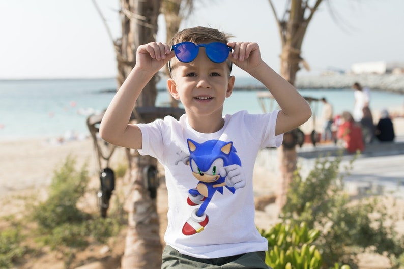 May include: A young child wearing a white t-shirt with a blue Sonic the Hedgehog graphic. The child is wearing blue sunglasses and is smiling.