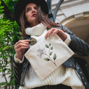May include: A cream-colored canvas pouch featuring a watercolor illustration of a lilac branch with purple flowers and green leaves. The pouch is held by a person wearing a black leather jacket and a white sweater.