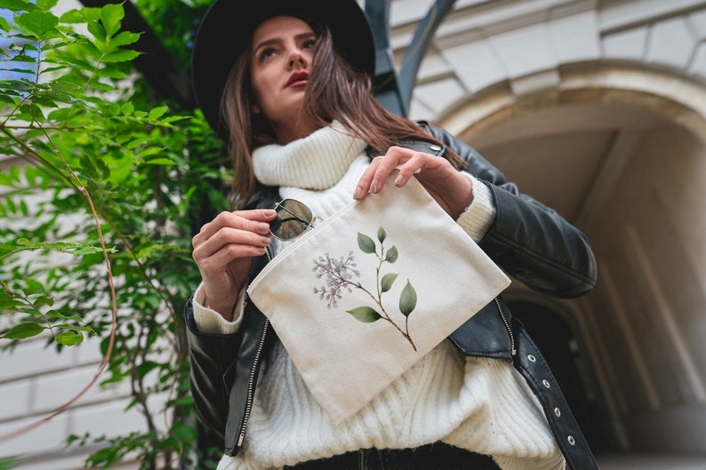 May include: A cream-colored canvas pouch featuring a watercolor illustration of a lilac branch with purple flowers and green leaves. The pouch is held by a person wearing a black leather jacket and a white sweater.