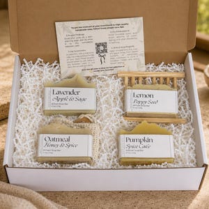 Op de afbeelding: Een kartonnen geschenkdoos met vier rechthoekige ambachtelijke zeepstukken met de labels Lavender Apple & Sage, Lemon Poppy Seed, Oatmeal Honey & Spice en Pumpkin Spice Cake, inclusief een instructiekaart en een houten zeepbakje.
