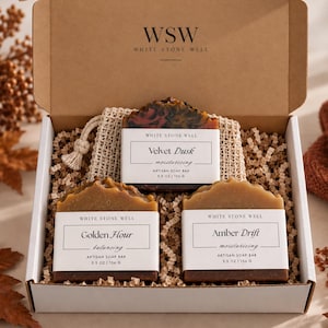 May include: A cardboard gift box containing three artisan soap bars labeled White Stone Well. The bars are named Velvet Dusk, Golden Hour, and Amber Drift, each weighing 5.5 oz. The soaps are nestled in paper filler with a small mesh bag included.