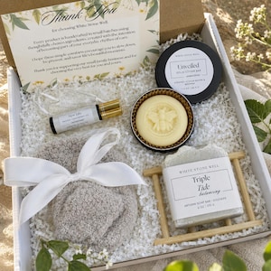 Op de afbeelding: Een geschenkdoos met een bedankkaart, een rollerflesje New Life zalfolie, een ronde vaste lotionbar met een bijenontwerp, grijze pluizige sokken met een wit lint en een stuk Triple Tide zeep op een houten zeepbakje.