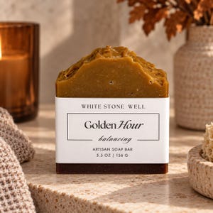 May include: A rectangular artisan soap bar with a textured, golden-brown top and a dark brown base. The white paper label reads: WHITE STONE WELL, Golden Hour, balancing, ARTISAN SOAP BAR, 5.5 OZ | 156 G, enclosed in a thin black rectangular border.