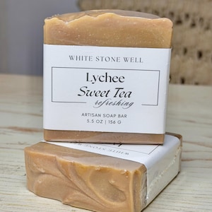 Puede incluir: Dos barras de jabón artesanal, una apilada encima de la otra. La barra superior tiene una etiqueta blanca que dice "White Stone Well" y "Lychee Sweet Tea refreshing". El jabón pesa 156 g. El jabón es de color marrón claro.