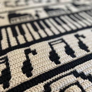 May include: Close-up of a black and white crochet blanket with a piano keyboard and musical notes pattern. The blanket is made with a textured stitch and has a subtle sheen.