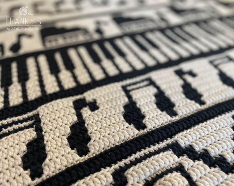 May include: Close-up of a black and white crochet blanket with a piano keyboard and musical notes pattern. The blanket is made with a textured stitch and has a subtle sheen.