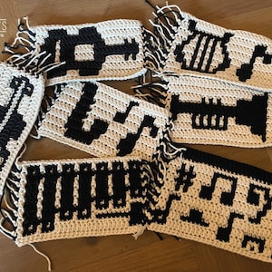 May include: Crochet patterns for musical notes, a trumpet, and a treble clef. The patterns are made with white yarn and black yarn. The patterns are fringed on the edges.