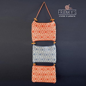 Op de afbeelding: Drie gehaakte wandhangers met geometrische patronen. De bovenste en onderste hangers zijn oranje en wit, en de middelste hanger is blauw en wit. De hangers zijn bevestigd aan een houten stok en opgehangen aan een touw.