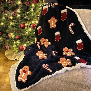 Op de afbeelding: Een donkerblauwe gehaakte deken met een witte rand heeft een feestelijk kerstontwerp. De deken is versierd met gingerbread men, snoepstokken en kousen.