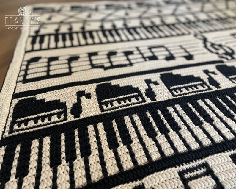 May include: Black and white crochet blanket with a musical theme. The blanket features a piano keyboard, musical notes, and a treble clef.