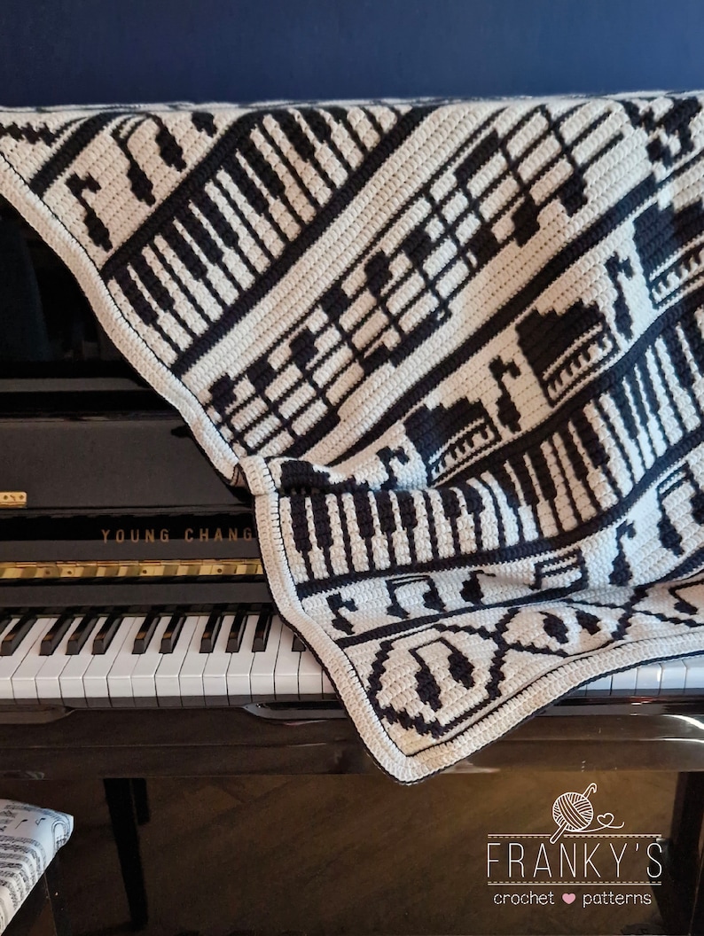 May include: A black and white crocheted blanket with a musical theme. The blanket features piano keys, treble clefs, and musical notes. The blanket is draped over a black piano with the words "YOUNG CHANG" on the front.