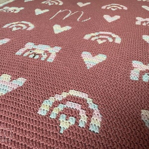 Op de afbeelding: Een roze gehaakte babydeken met regenboog-, hart- en dierenmotieven. De deken heeft de letter "M" in het midden.