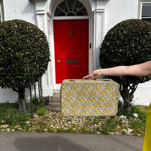 May include: A vintage floral patterned suitcase with a yellow, pink, and green design. The suitcase is being held by a hand in front of a red door.