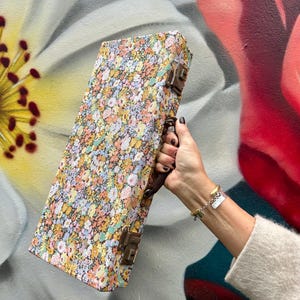 May include: A rectangular case covered in a floral pattern, with a brown handle and metal clasps. The case displays a colourful pattern of small flowers in pink, yellow, and blue. It is held against a painted floral background.