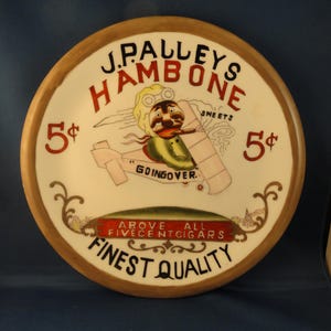 May include: A white ceramic plate with a brown rim features a vintage advertisement for J. Palley's Hambone cigarettes. The ad features a cartoon illustration of a pilot in a plane with the text "Going Over" and the slogan "Above All Five Cent Cigars Finest Quality". The plate is decorated with a red and white border and the price "5 cents" is written in red on each side.