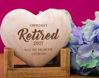 May include: White heart-shaped stone with the words "Officially Retired 2021 Not My Problem Anymore" engraved on it. The stone is sitting on a wooden stand and a blue and green flower is in the background.