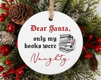 May include: A white, round Christmas ornament with the text "Dear Santa, only my books were Naughty" in black and red script. A stack of books is illustrated next to the text. The ornament hangs from a jute string and is surrounded by greenery, pinecones, and red berries.
