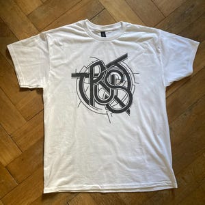Könnte beinhalten: Ein weißes T-Shirt mit kurzen Ärmeln und einem großen schwarzen Grafikdruck auf der Brust. Das Design besteht aus dem Wort TRUST, das mit fetten, sich überlappenden Linien und geometrischen Kreiselementen komplex dargestellt ist.