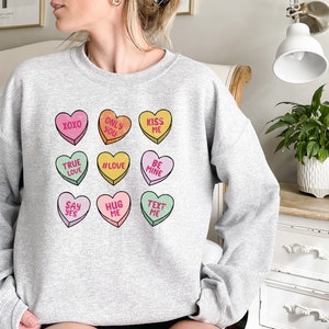 Pu&ograve; includere: Felpa grigia con una grafica di nove cuori di caramelle colorati con diversi messaggi, tra cui "XOXO", "Only You", "Kiss Me", "True Love", "#Love", "Be Mine", "Say Yes", "Hug Me" e "Text Me".
