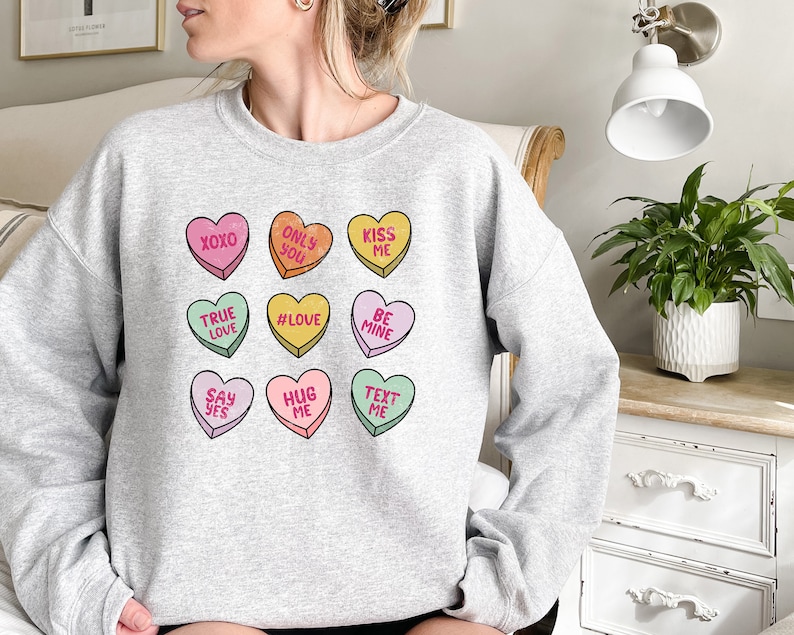 Pu&ograve; includere: Felpa grigia con una grafica di nove cuori di caramelle colorati con diversi messaggi, tra cui "XOXO", "Only You", "Kiss Me", "True Love", "#Love", "Be Mine", "Say Yes", "Hug Me" e "Text Me".
