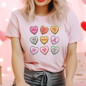 Pu&ograve; includere: Maglietta rosa con una grafica di nove cuori di caramelle con messaggi diversi, tra cui "XOXO", "True Love", "#Love", "Hug Me", "Say Yes", "Only You", "Kiss Me", "Be Mine" e "Text Me".