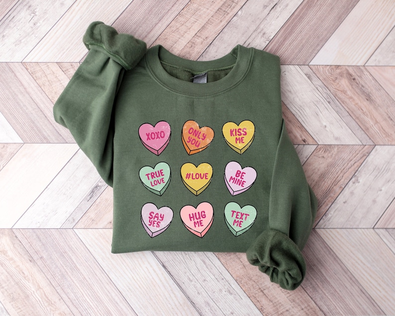 Pu&ograve; includere: Una felpa verde a tema San Valentino. Presenta un motivo di nove cuori con diverse frasi, tra cui "XOXO", "True Love", "#Love", "Only You", "Kiss Me", "Be Mine", "Say Yes", "Hug Me" e "Text Me".