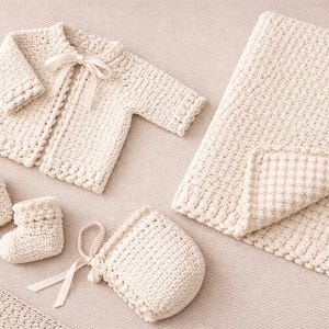 Può includere: Un set per neonati lavorato a mano, color crema, composto da una giacca con fiocco, una coperta, un cappellino con fiocco e un paio di scarpine. Il set è realizzato in filato morbido e strutturato.
