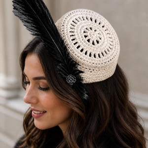 May include: A cream-colored crochet hat with a circular design, adorned with a large black feather and a small, decorative black embellishment. The hat is a unique accessory, perfect for special occasions.
