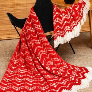 May include: A red and white crocheted blanket with a chevron pattern and white fringe. The blanket is draped over a black chair and a small table. The blanket's design features a repeating zigzag pattern.