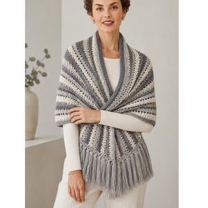 May include: A gray, white, and cream-colored crochet shawl with a striped pattern and fringe. The shawl is draped over the shoulders. The shawl is made of soft yarn and is a cozy accessory.