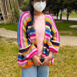 May include: A colorful, striped crochet cardigan in shades of pink, orange, blue, and green. The open-front sweater is worn over a pink top and paired with light blue jeans. The outfit is completed with white sneakers.