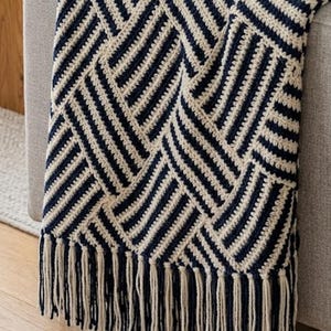 May include: A navy blue and cream-colored crocheted throw blanket with a geometric pattern and fringe detail. The blanket is draped over the arm of a gray sofa. The room has a neutral color palette.