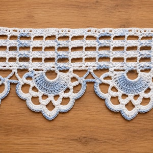 Puede incluir: Un borde de encaje de ganchillo blanco y azul claro con un patrón de rejilla rectangular repetitivo en el borde superior y una serie de motivos festoneados en forma de abanico en el borde inferior, acabado con delicados bucles decorativos calados.