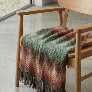 Puede incluir: Una manta con estampado de chevrón multicolor con flecos, colocada sobre una silla de madera. La manta presenta tonos de verde, marrón y naranja. La silla es de madera clara.