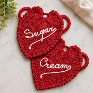 Pode incluir: Duas pegas de croché vermelhas em forma de coração com bordas recortadas e pegas integradas. Uma pega apresenta o texto bordado a branco "Sugar" e a outra o texto bordado a branco "Cream" numa fonte de escrita cursiva.