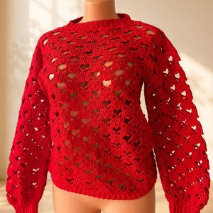 Puede incluir: Un suéter rojo brillante de manga larga con un patrón de ganchillo abierto que presenta recortes repetitivos en forma de corazón en todo el cuerpo y las mangas. La prenda tiene un cuello redondo y puños acanalados en las muñecas y el dobladillo.