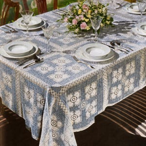 May include: A rectangular tablecloth featuring a blue and white grid pattern with intricate lace floral motifs. The table is set with white ceramic plates, clear glass goblets, silver forks and spoons, and a centerpiece of pink and yellow flowers.