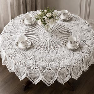 Può includere: Una tovaglia rotonda in uncinetto bianco caratterizzata da un intricato motivo radiale di elementi ripetuti a forma di foglia. La tovaglia è stesa su un tavolo circolare ed è apparecchiata con quattro tazze da tè in ceramica bianca.