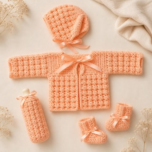May include: A peach-colored crochet baby set, including a jacket, bonnet, bottle cover, and booties. Each piece is adorned with a satin ribbon bow. The set is made of a textured stitch, perfect for a newborn.