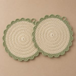 Op de afbeelding: Twee crèmekleurige, ronde gehaakte pannenlappen met een geschulpte saliegroene rand. Elke pannenlap heeft een kleine lus om op te hangen. Het haakpatroon is een spiraalvormig ontwerp, waardoor een gestructureerd oppervlak ontstaat. Handgemaakte keukenartikelen.