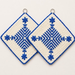May include: Two square crochet potholders in cream with a vibrant blue geometric snowflake pattern. Each potholder features a central star motif, radiating lines, and a small silver metal ring at the top corner for hanging, showcasing detailed needlework.