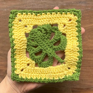 Könnte beinhalten: Ein gehäkeltes Quadrat mit einem grünen Blattdesign in der Mitte. Das Quadrat ist von einem gelben Rand und einem grünen Außenrand umgeben.